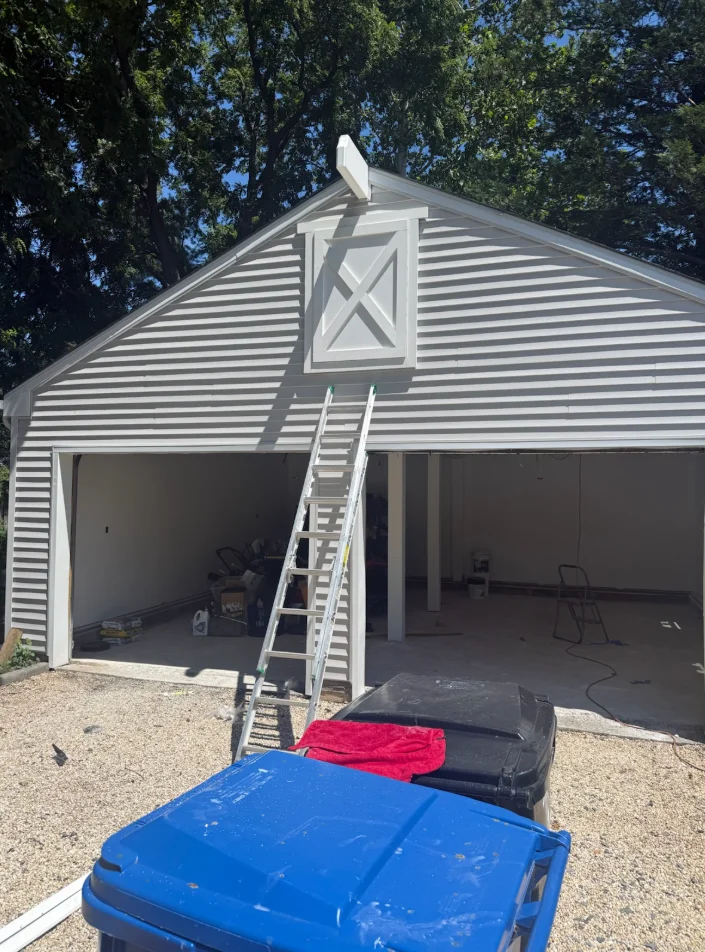 Two-car garage with gray and white horizontal siding, peaked roof, and X-pattern gable window