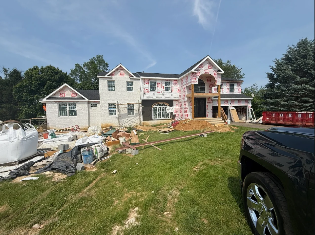 Two-story home under construction with white brick, gabled roofs, and house wrap during framing phase