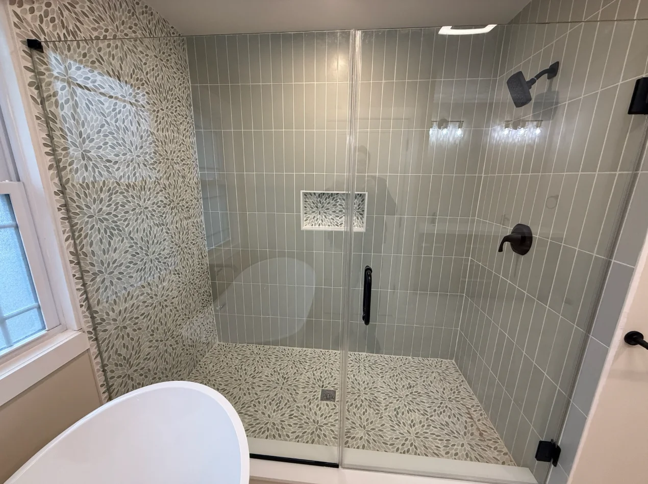 Frameless glass shower enclosure with geometric mosaic tile, vertical subway tile walls, and black fixtures